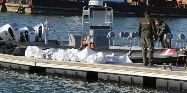 Mehrere Leichensäcke liegen auf einem Bootssteg, daneben zwei tunesische Grenzschützer, die sich an einem Boot zu schaffen machen