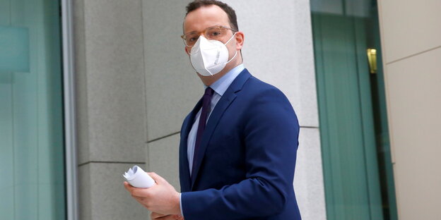 Jens Spahn steht vor einem Gebäude, trägt FFP2-Maske und hält ein gerolltes Blatt in den Händen.