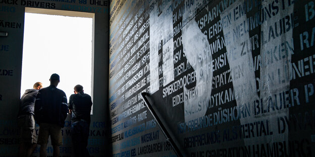 Am Ende einer im Dunkeln liegenden Treppe stehen Menschen an einer in gleißendes Licht getauchten Maueröffnung. Auf den Wänden stehen die Namen von HSV-Fanclubs und in großen Lettern "HSV"