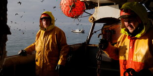 Zwei Männer stehen auf einem Boot und blicken in die Kamera. Im Hintergrund ist das Meer.