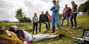 Eine Leiche liegt auf einer Wiese, an einen Stein gelehnt. Davor stehen ErmittlerInnen, rechts ein medizinischer Koffer, im Hintergrund ein Holzhaus