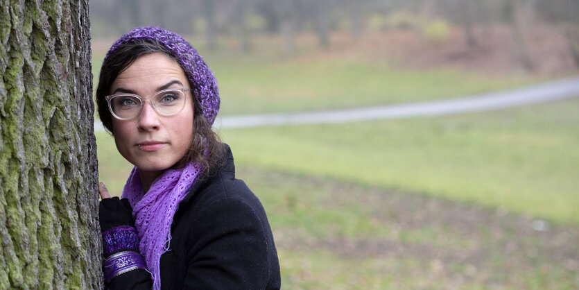 Die Musikerin Sookee lehnt an einem Baum auf einer Wiese. Sie trägt eine Brille mit einer durchsichtigen Fassung und hat die dunklen langen Haare unter einer lila Mütze versteckt.