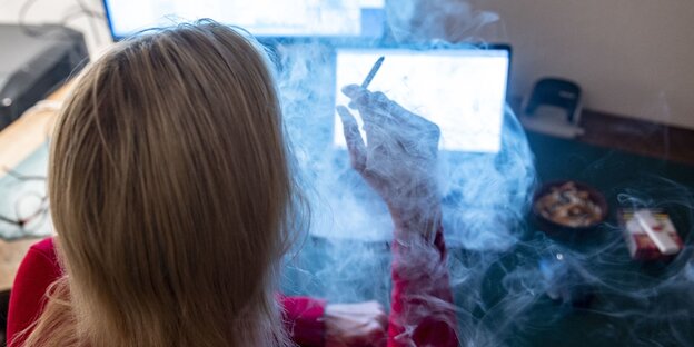 Das Bild zeigt eine blonde Frau von hinten. Sie sitzt vor einem Computer und raucht. Daneben stehen volle Aschenbecher.