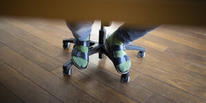 Blick unter den Schreibtisch im Home Office: Ein Mann trägt grüne Socken und Hausschuh-Sandalen