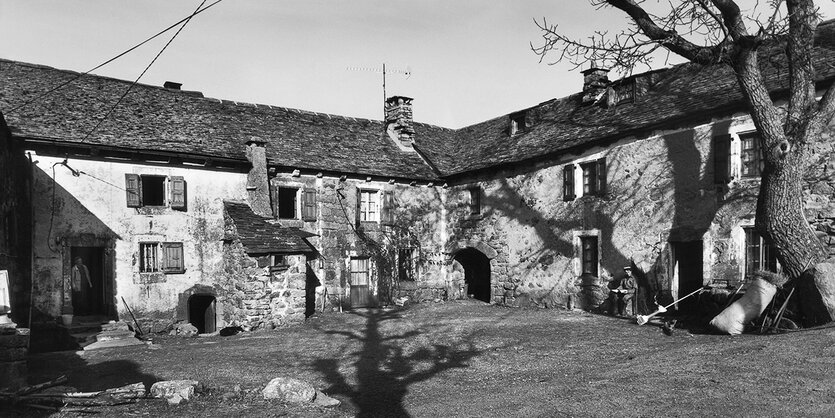 Schwarzweißaufnahme eines Bauernhofs im Winter. Der Schatten eines Baums fällt in den Hof