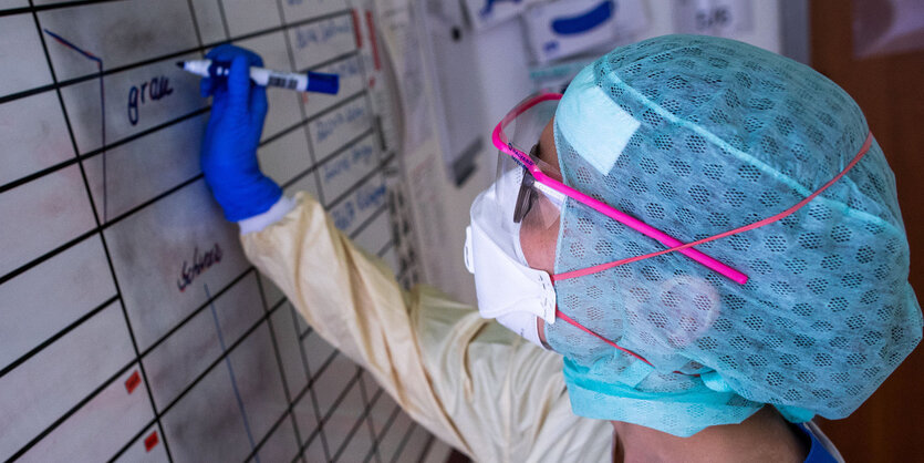 Eine Krankenschwester mit Schutzausrüstung, das heißt Overall, Handschuhe, Schutzbrille, FFP Maske und Haube, trägt auf einer Tafel die Belegung der Betten ein.