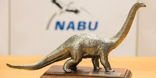 Eine kleine messigfarbende Dinosaurier Figur steht auf einem Holzsockel