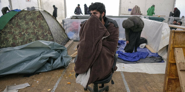 Ein Migrant sitzt mit einer Decke zugedeckt auf einem Stuhl im Lager Lipa
