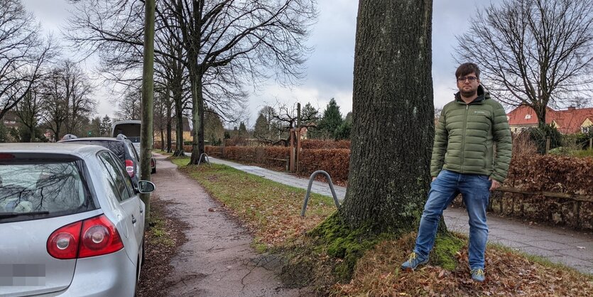 Ein Mann steht vor einem Baum, links davon ein schmaler Radweg und en parkendes Auto, rechts davon ein schmaler Gehweg