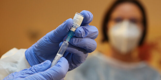 Ein Arzt zieht eine Spritze mit Impfstoff gegeb Covid-19 auf. Im Hintergrund eine Frau mit Mund-Nasen-Schutz