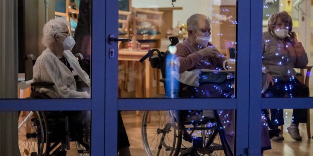 Drei Altenheim-BewohnerInnen sitzen mit Masken hinter einer verschlossenen Glastür