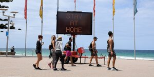 Stay at home, steht auf einer digitalen Anzeige, davor Menschen am Strand.