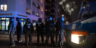 Polizisten stehen am Silvesterabend neben einem Einsatzfahrzeug auf dem Kottbusser Damm, im Hintergrund ein paar Raketen am Himmel