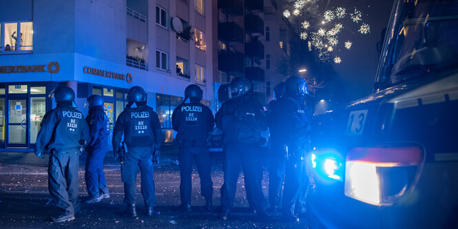 Polizist:innen stehen auf dem Kottbusser Damm und überwachen das Böllerverbot. Am Nachthimmel explodiert Feuerwerk.