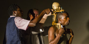 Drei Männer auf der Bühne, einer trägt ein Skelett auf der Schulter.