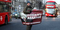 Mann auf einer Straße in London, man sieht zwei rote Doppeldeckerbusse. In den Händen hält er ein Transparent: "Hands off Assange"
