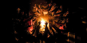 Viele Menschen sitzen nachts im Kreis um ein offenes Feuer und strecken ihre Hände in die Wärme