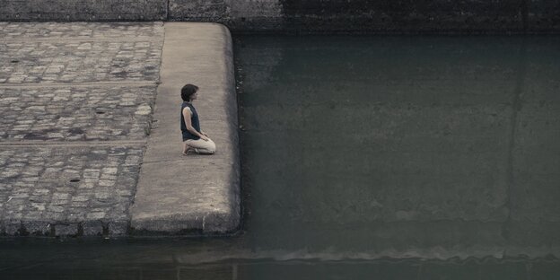 Auf dem gemauerten Ufer eines Gewässers kniet die Künstlerin Naomi Wiener