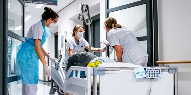 Drei Krankenhausmitarbeiterinnen hantieren an einem leeren Krankenbett.