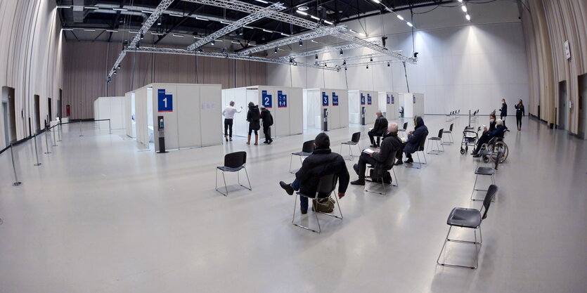 Menschen sitzen in einer Halle auf Stühlen, die weit voneinander weg stehen. Vor ihnen sind kleine Impfkabinen aufgebaut