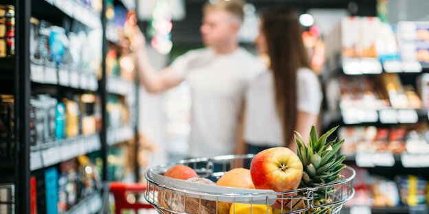 Einkaufskorb im Supermarkt mit Obst und zwei junge Menschen beim Einkaufen