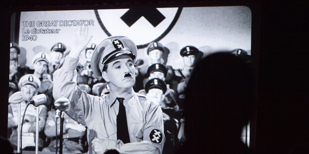 Ein Filmausschnitt aus "Der große Diktator", mit Charlie Chaplin als Hitler im Vordergrund. Im Hintergrund Nationalsozialisten und die Hakenkreuzflagge