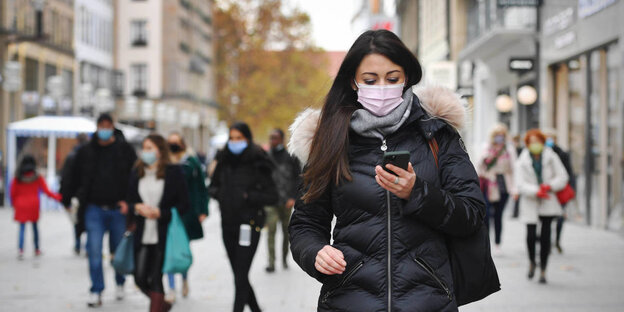 Eine Frau mit Mundnasenschutz blickt auf ihr Smartphone in einer Fußgängerzone