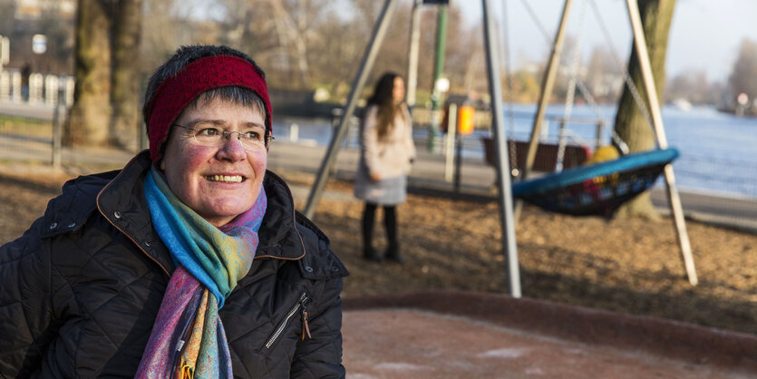 Helga Krull, Anfang 60, sitzt auf einem Spielplatz. Sie trägt einen bunten Schal und ein rotes Stirnband.