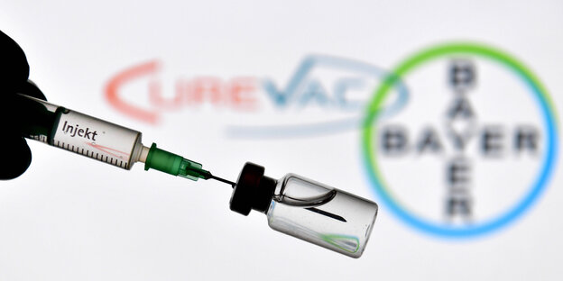 Eine Hand in Gummihandschuhe gehuellt haelt eine Einwegspritze und eine Impfdose vor den Logos von CureVac und Bayer