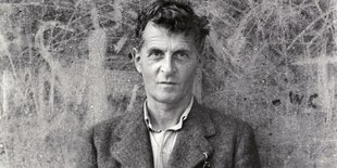 Ludwig Wittgenstein steht vor einer grauen Wand auf der Kratzer und Botschaften eingeritzt sind