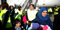 Eine Frau mit blauem Kopftuch und zwei Kindern steht lächelnd vor einem Flugzeug, dahinter Sicherheitsleute