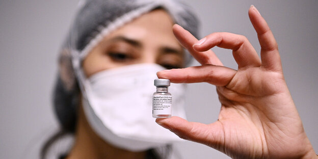 Eine Frau mit Mundschutz und Haube hält eine Ampulle Impfstoff zwischen Daumen und Zeigefinger