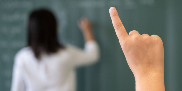 Ein Schüler meldet sich während einer Unterrichtsstunde während die Lehrerin an die Tafel schreibt.