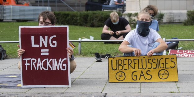 Junge Menschen sitzen mit Maske und Protestschildern auf dem Boden und demonstrieren gegen Flüssiggas. Auf den Schildern steht LNG=Fracking, auf einem anderen "Überflüssiges Gas"