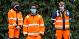 Drei Männer in orangenen Overalls stehen vor einem grünen Busch, sie tragen alle Maske