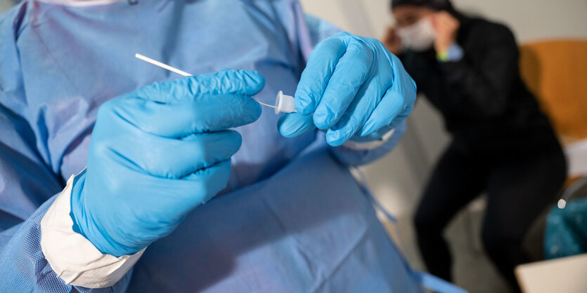 Eine Person in Schutzkleidung steckt ein Stäbchen von einem coronatest in eine Röhre. Im Hintergrund sitzt eine Person, die sich gerade die Maske wieder aufsetzt.