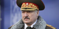 Lukaschenko in einem Militärmantel mit riesigem Militärgut. Der Machthaber von belarus schaut grimmig. Er hat einen Bart.