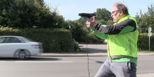 Ein Mann mit einem Föhn in der Hand zielt auf ein Auto, das vorbeifährt