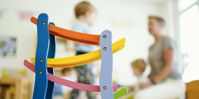 Man sieht ein buntes Holzspielzeug, im Hintergrund ein Kleinkind und eine Erzieherin