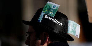 Ein Demonstrant mit einem Hut, auf dem BlackRock steht mit zwei 100-Euro Banknoten