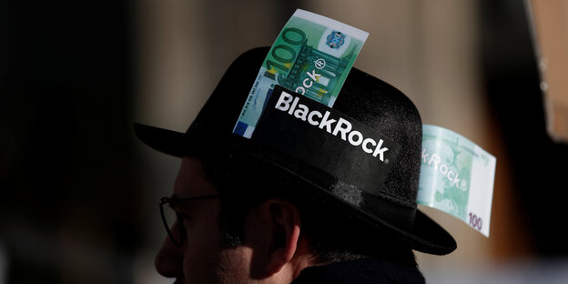 Ein Demonstrant mit einem Hut, auf dem BlackRock steht mit zwei 100-Euro Banknoten