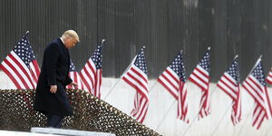 Donald Trump läuft Treppen vor der US-Mexikanischen Mauer hinunter - im Hintergrund US-Flaggen