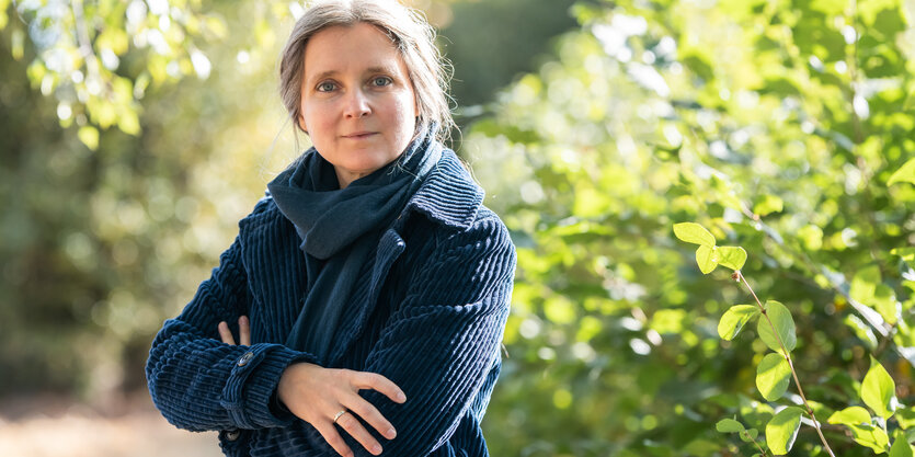 Marion Poschmann steht in blauem Mantel vor grünendem Buschwerk