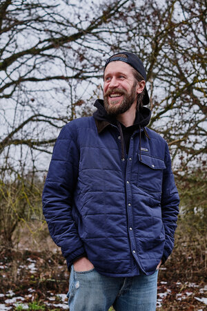 Ein Mann in blauer Stzeppjacke und mit einer Kappe auf dem Kopf steht vor einer Hecke und lacht Ein Mann in blauer Stzeppjacke und mit einer Kappe auf dem Kopf steht vor einer Hecke und lacht