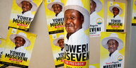 Plakate mit Profil von Yoweri Museveni auf gelbem Hintergrund. Sie kleben an einem Pfahl im Vordergrund und an Händen im Hintergrund