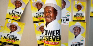 Plakate mit Profil von Yoweri Museveni auf gelbem Hintergrund. Sie kleben an einem Pfahl im Vordergrund und an Händen im Hintergrund