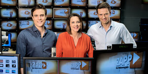 Die drei Zapp-Moderator*innen Constantin Schreiber, Kathrin Drehkopf und Johannes Jolmes nebeneinander vor einer Fernsehwand