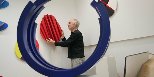 Der Künstler Micheal M. Heyers hängt eine rote abstrakte Plastik an die Wand, im Vordergrund ist eine große blaue Kreisform zu sehen
