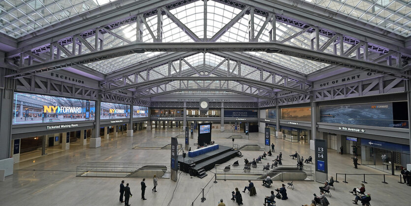 Eine große lichtdurchflutete Halle, in der sich sehr wenige Menschen aufhalten. Sie sitzen auf Stühlenund sind die teilnehmer der Eröffnungszeremonie für die Moynihan Train Hall