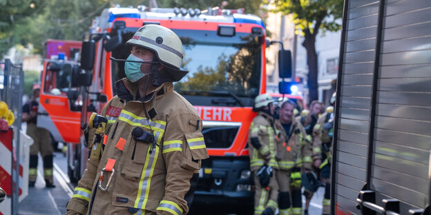 Ein Feuerwehrmann mit Mundschutz an einem Löschfahrzeug neben welchem weitere Kollegen stehen